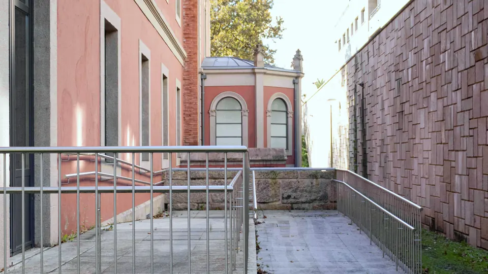 Rampa de acceso con barandillas de acero entre un edificio histórico de fachada rosada y un muro de aplacado de piedra