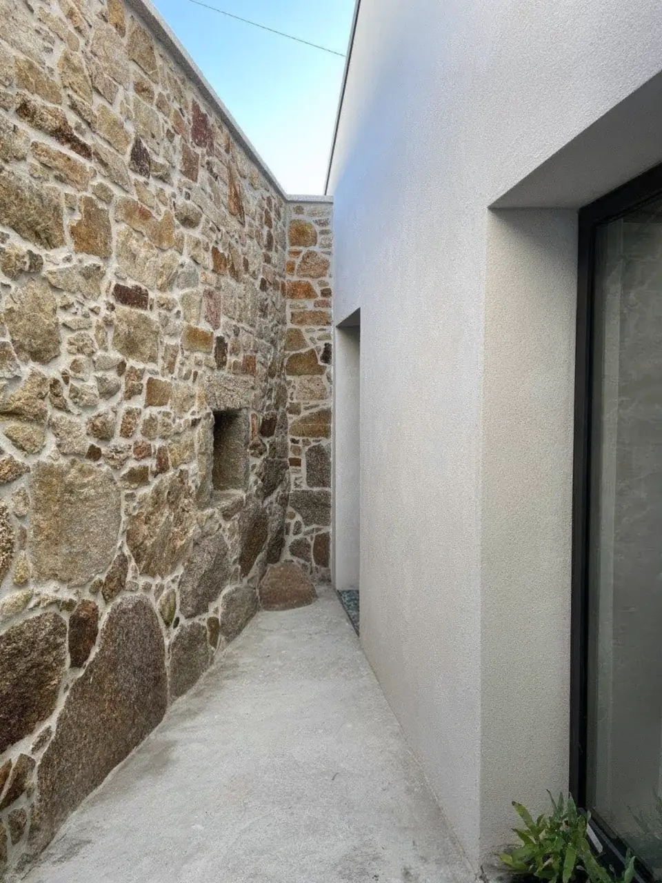Pasillo exterior estrecho entre un muro antiguo de mampostería de piedra y una fachada enfoscada en blanco con un gran ventanal